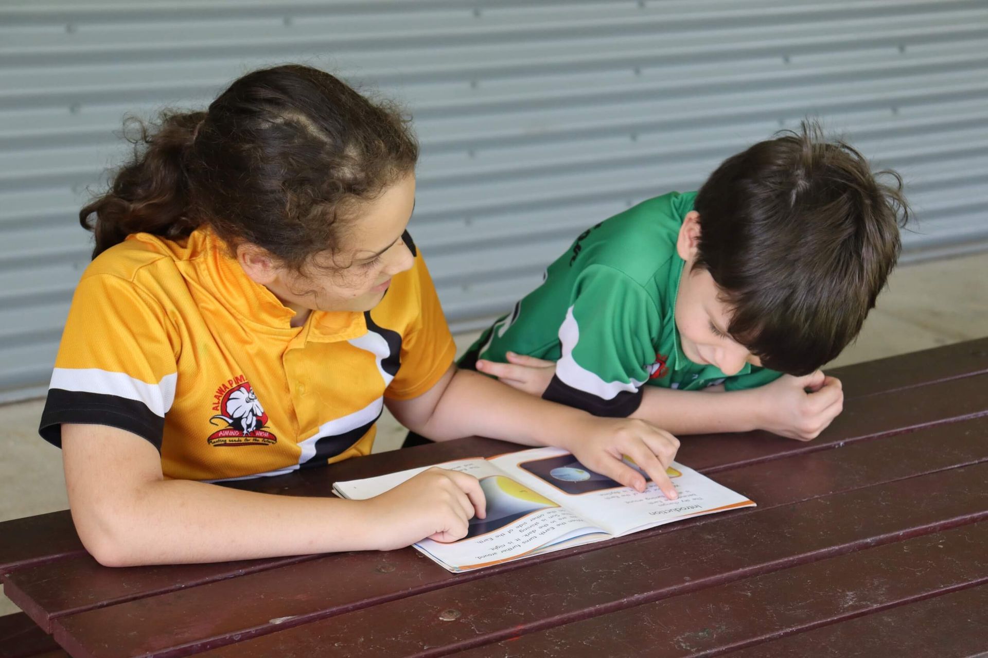 Two children reading a book together at a wooden table. One in yellow shirt points at the page; the other in green looks down.