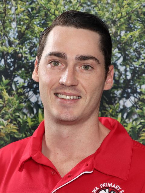 Smiling man in red polo shirt, WA Primary School emblem, outdoors with green foliage background.