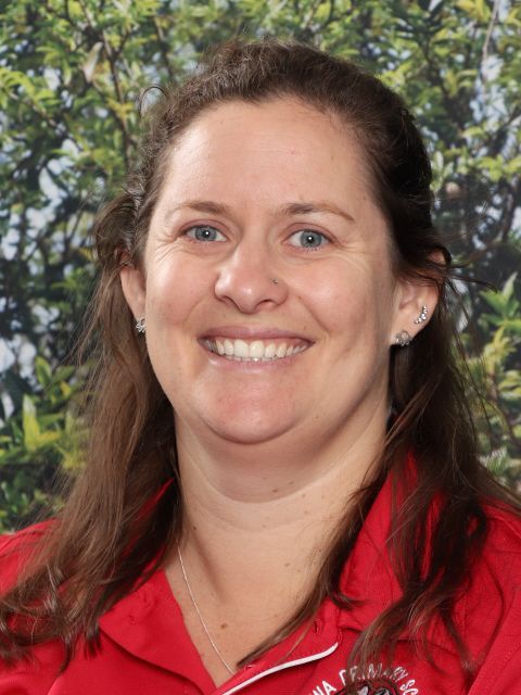 Woman with fair skin smiles, wearing a red shirt. Background is blurred greenery.