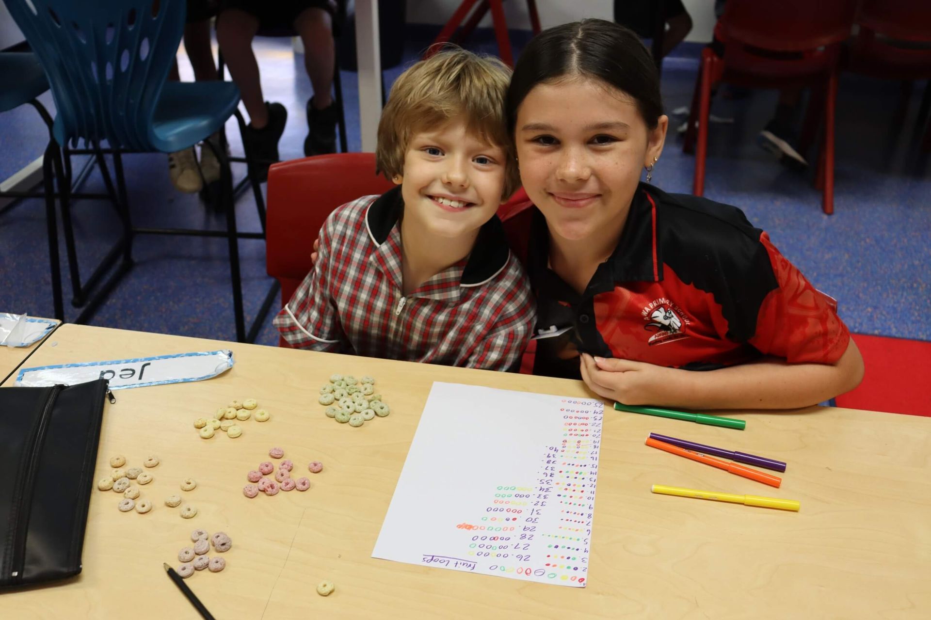 Two children smiling at a table with colorful cereal and a paper with patterns.