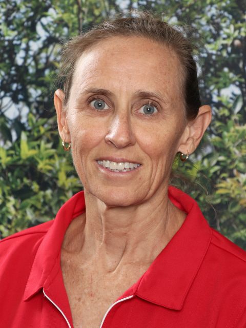 Woman with fair skin smiles, wearing a red shirt, in front of blurred greenery.