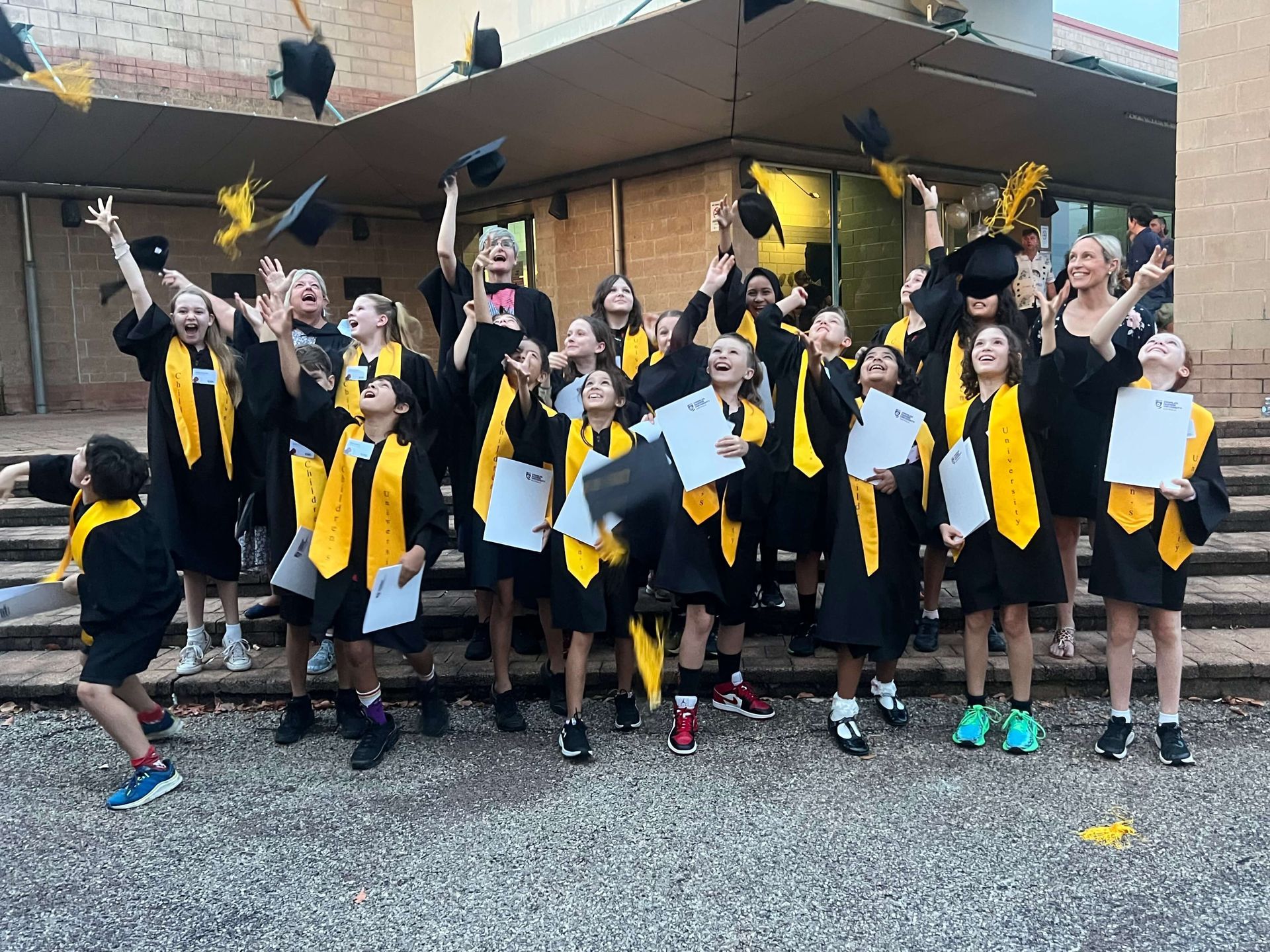 Graduating students in black gowns and yellow stoles throw caps in the air outside.