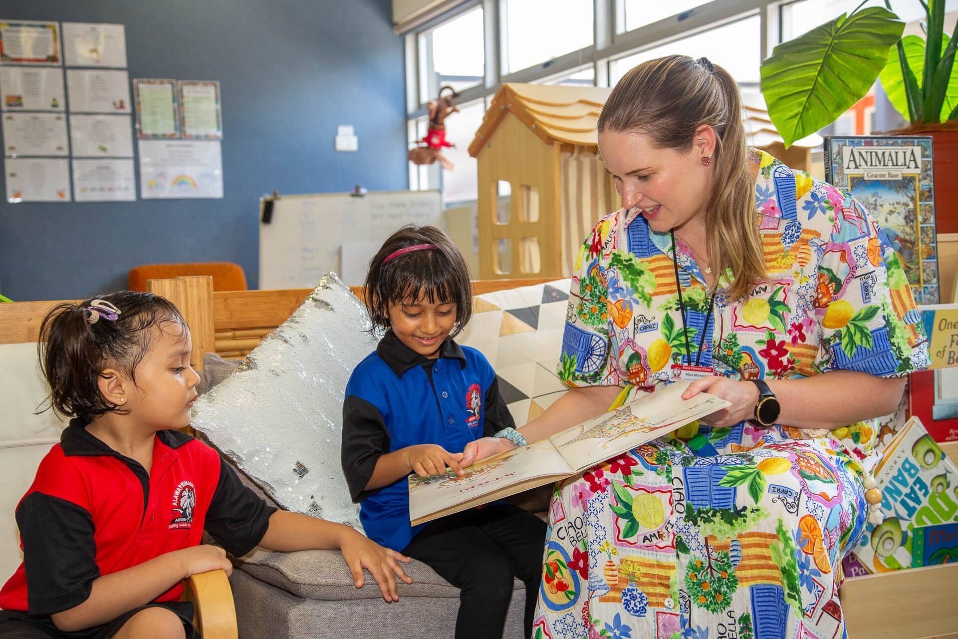Teacher reads a book to two young students in a colorful classroom setting with a playhouse and library shelves.