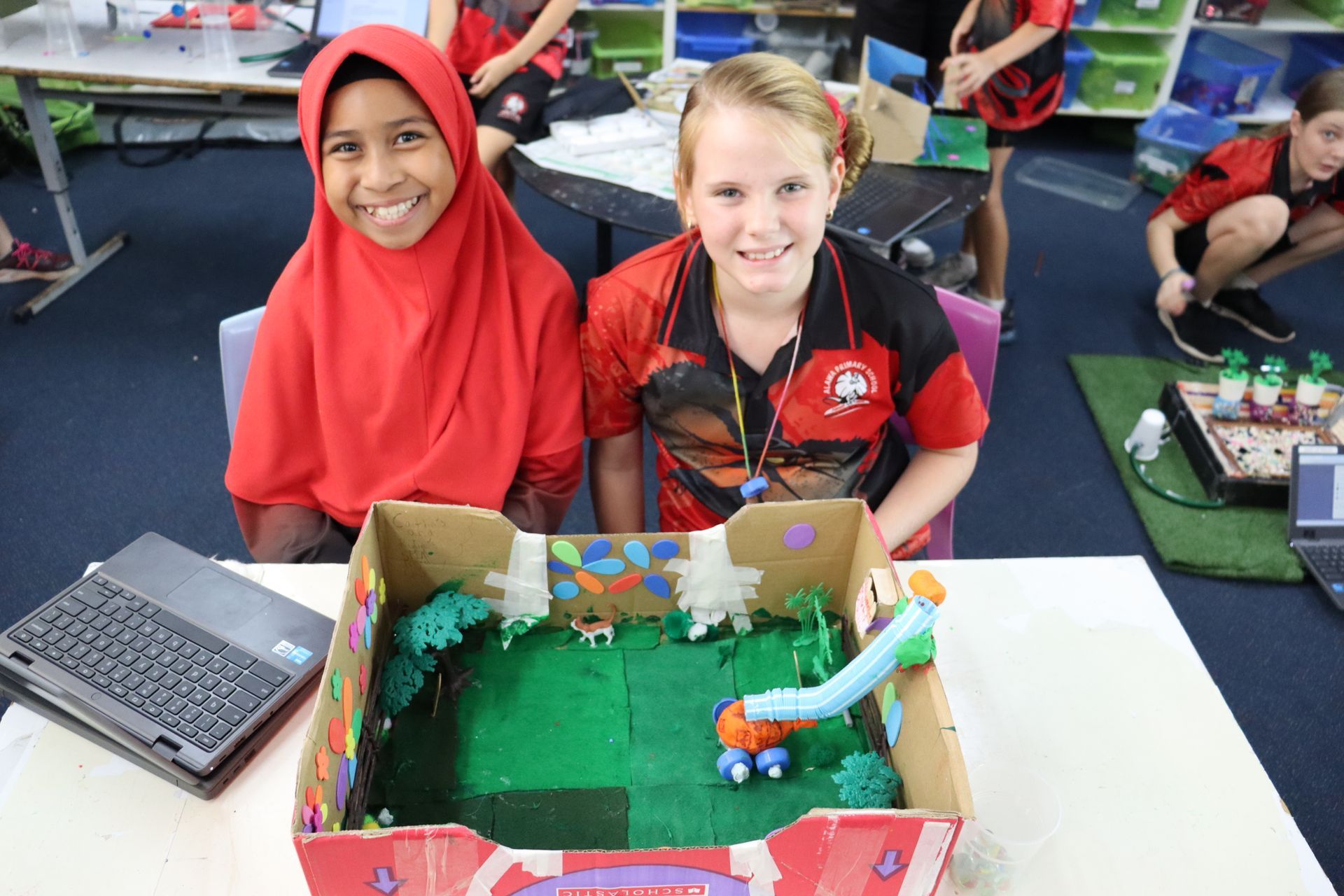 Two girls smiling, presenting a cardboard box diorama with a slide, greenery, and small figures on a table in a classroom.