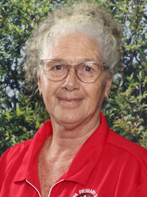 Woman with glasses and curly white hair, wearing a red polo shirt, smiles in front of a green backdrop.