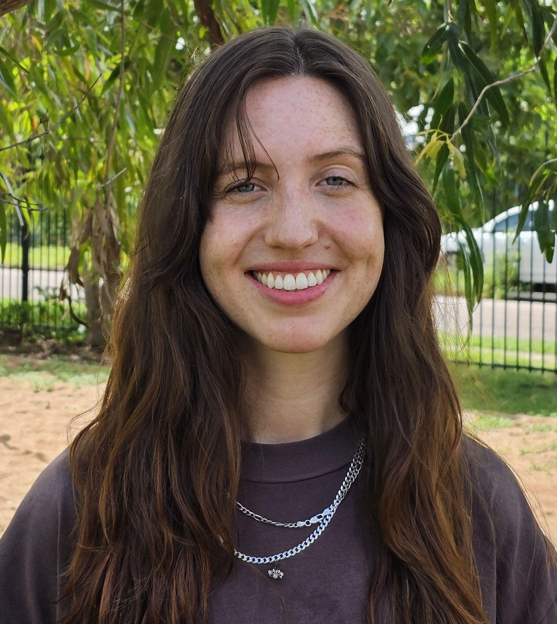 Smiling man in red polo shirt, WA Primary School emblem, outdoors with green foliage background.