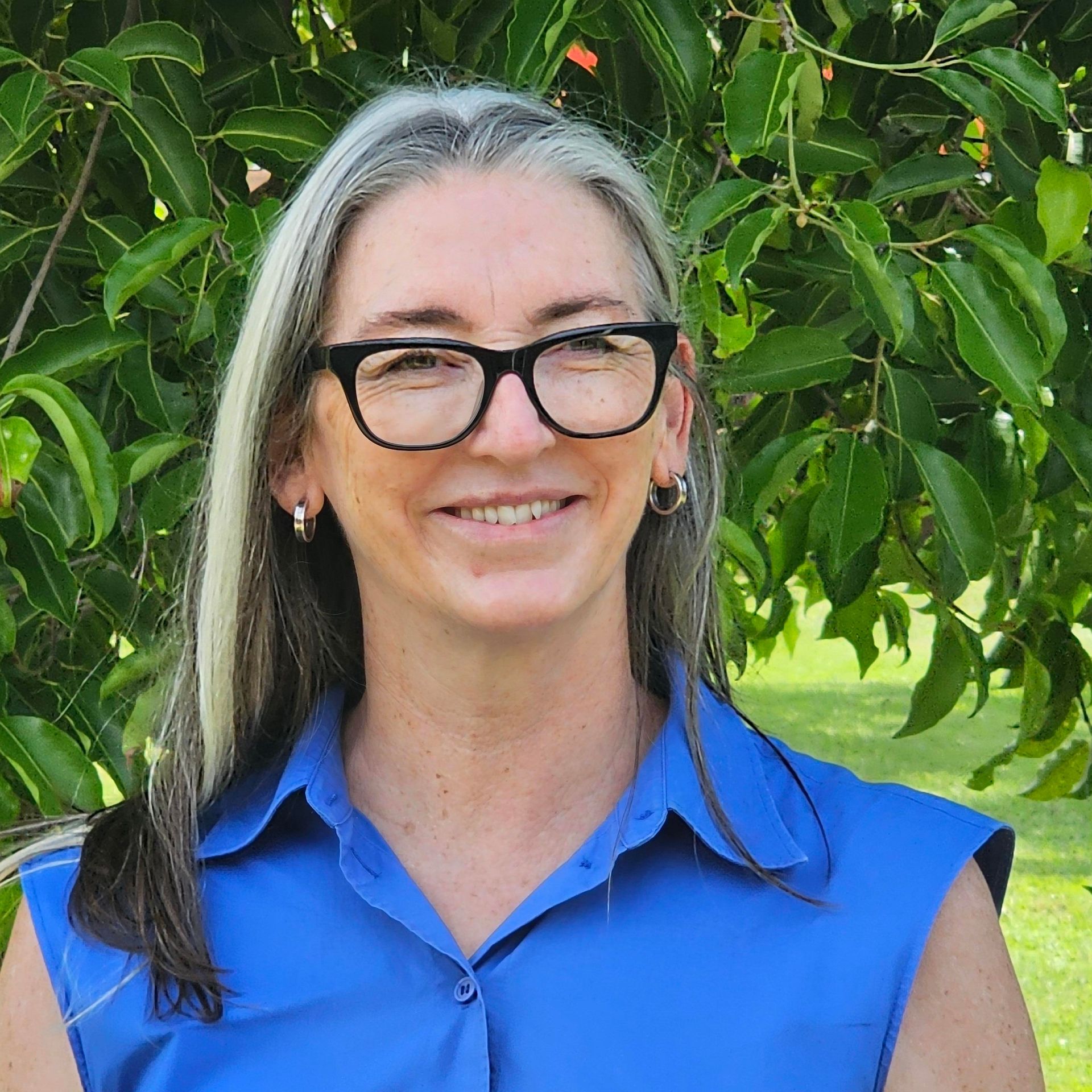 Woman with fair skin smiles, wearing a red shirt. Background is blurred greenery.