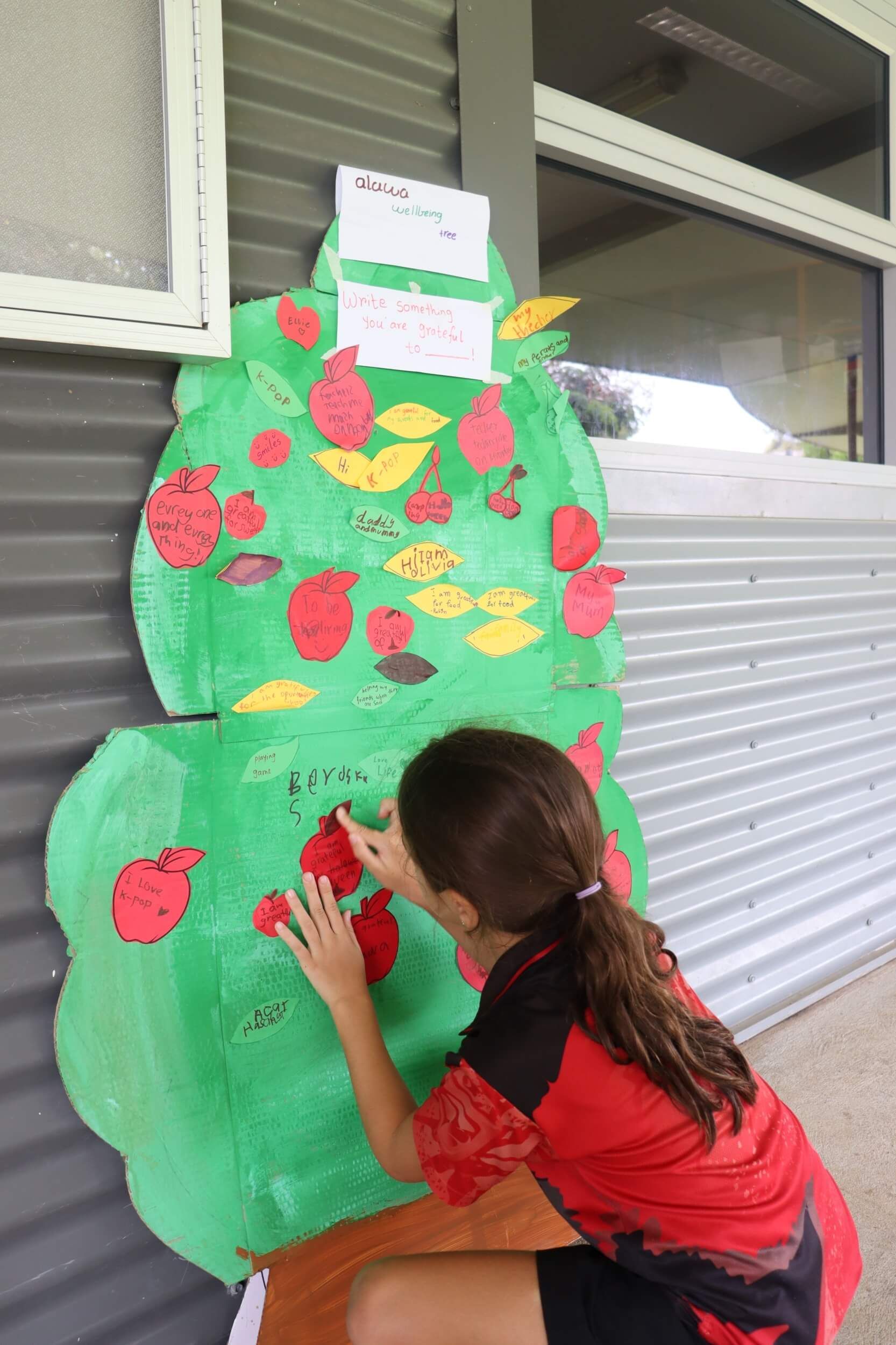 Girl adding a paper apple to a cardboard tree with notes attached. Outdoors, near a building.