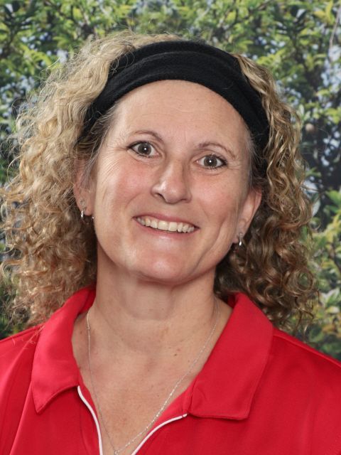 Woman with curly blonde hair and black headband smiles in a red shirt against a green backdrop.