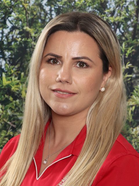 Woman with blonde hair, wearing a red shirt, smiling at the camera with a background of green foliage.