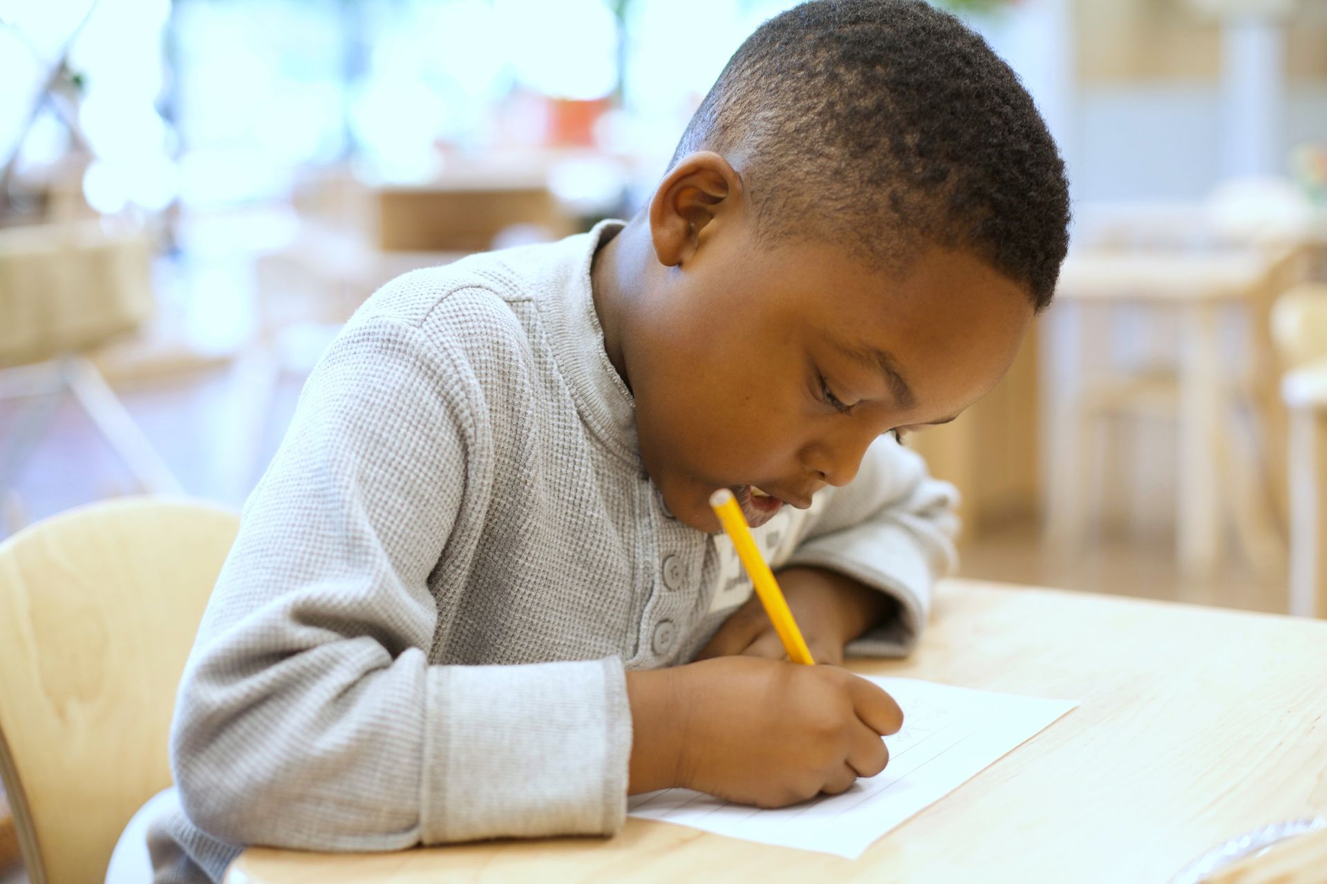 Child writing with a pencil at a table in a bright classroom