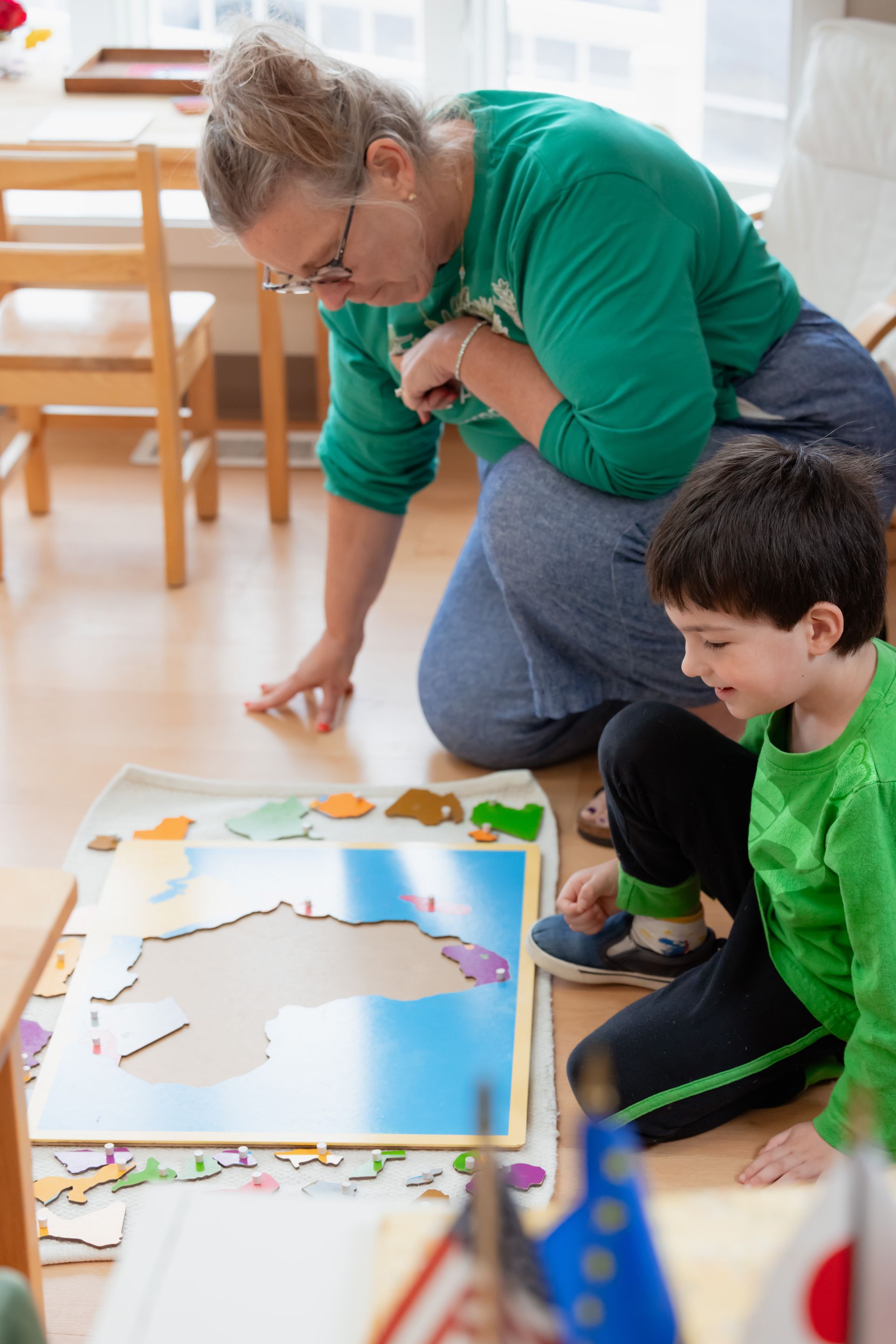 Two people kneel on a floor, working together on a colorful puzzle in a bright room.