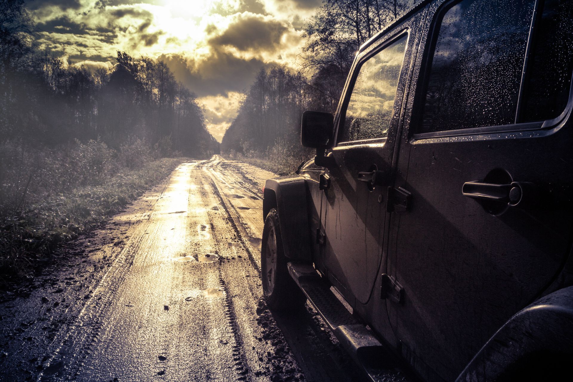 Jeep on a muddy road, trees on either side, sun shining through the clouds.