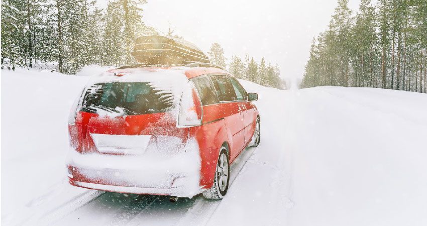 Red minivan driving on a snowy road in a wintery forest setting.
