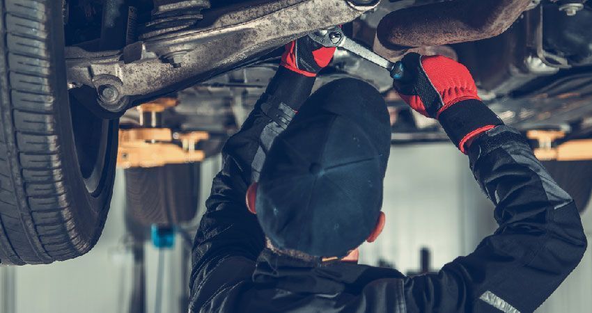 Mechanic working under a car, using a wrench. Wearing black cap, gloves and overalls, in a garage.