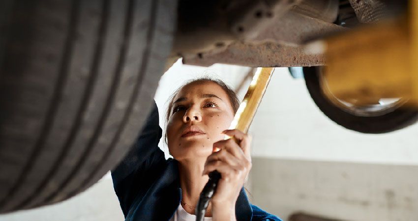 Woman in garage inspecting car underside with a light, looking up with focused expression.