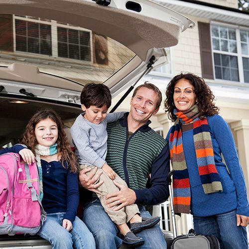 Family of four smiles at camera while loading car trunk in front of their house.