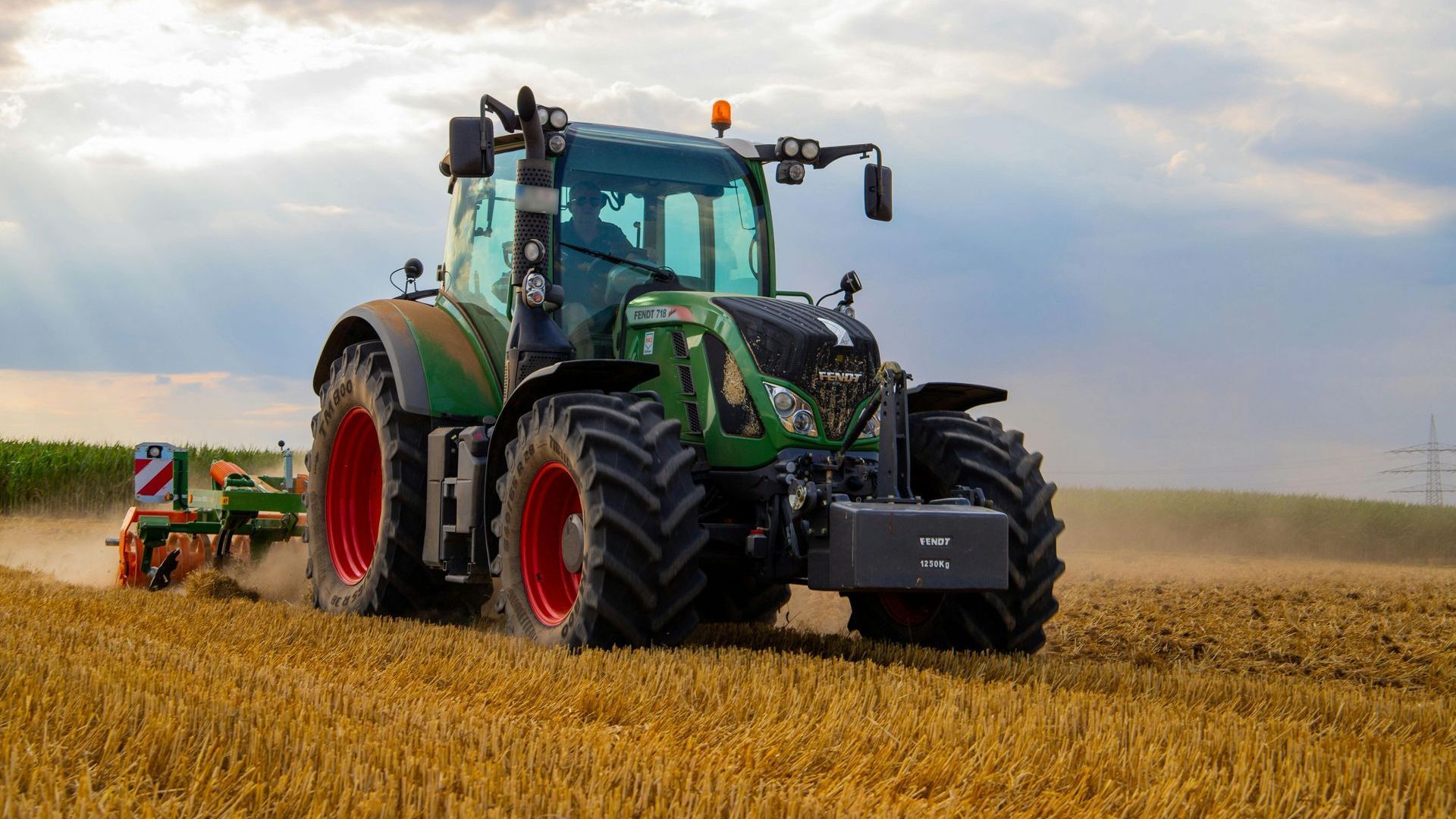 Green tractor plowing a wheat field under a cloudy sky.