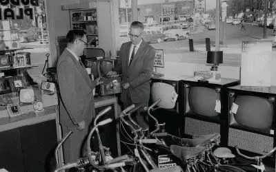 Two men in suits looking at a TV in a shop, bikes in foreground, 1960s.