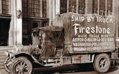 Vintage Firestone truck with canvas covering, parked in front of a building. Text on the side says