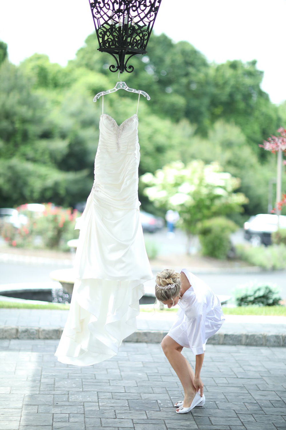 A bride is putting on her shoes in front of a wedding dress hanging on a hanger.