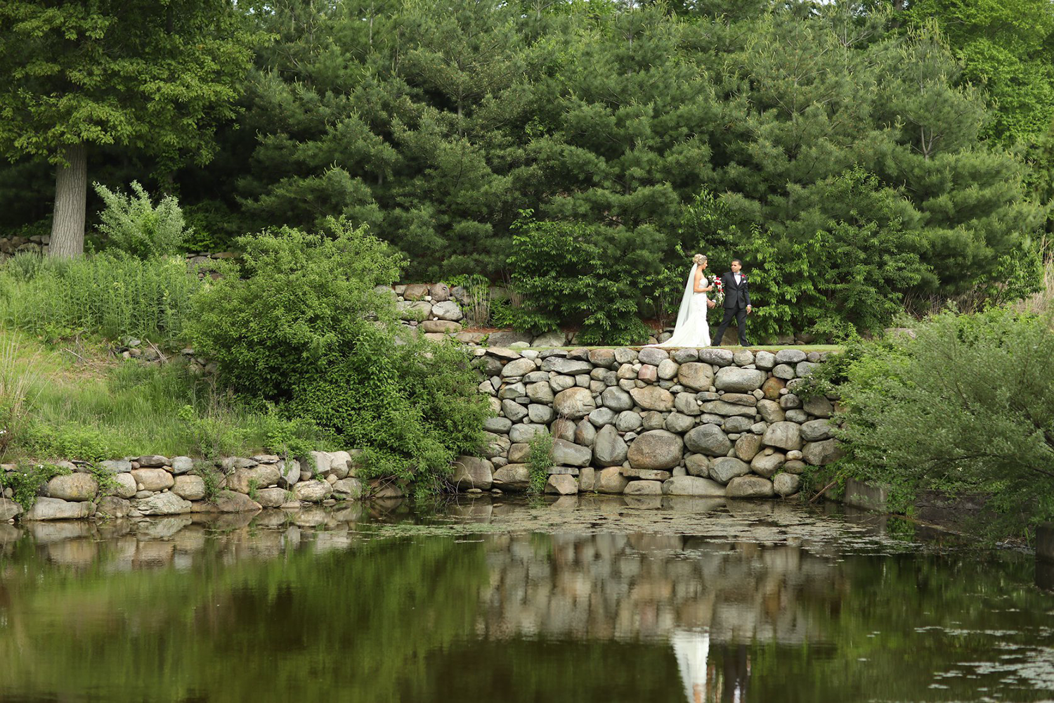 A bride and groom are standing on a stone bridge over a pond.