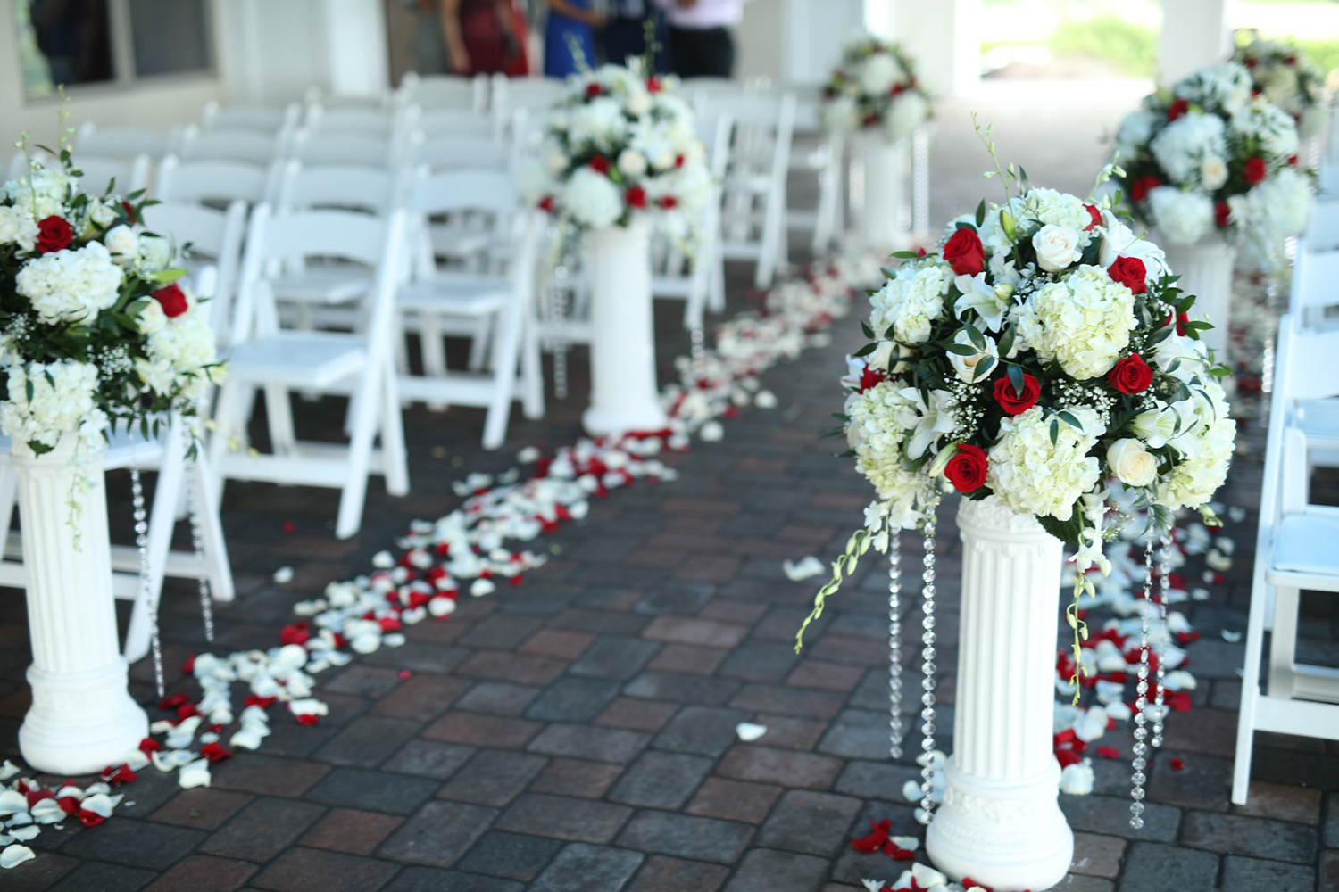 A row of white chairs are lined up along a brick walkway decorated with red and white flowers.