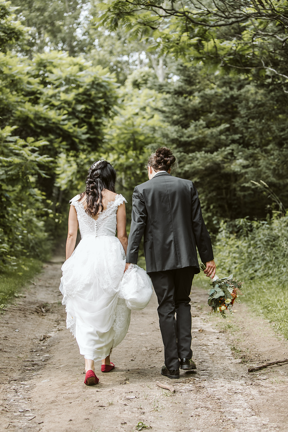 A bride and groom are walking down a dirt road holding hands.