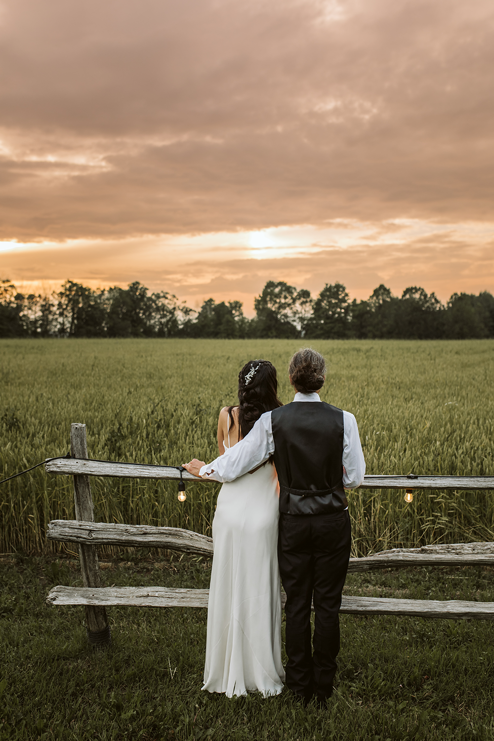 A bride and groom are standing next to a wooden fence in a field at sunset.