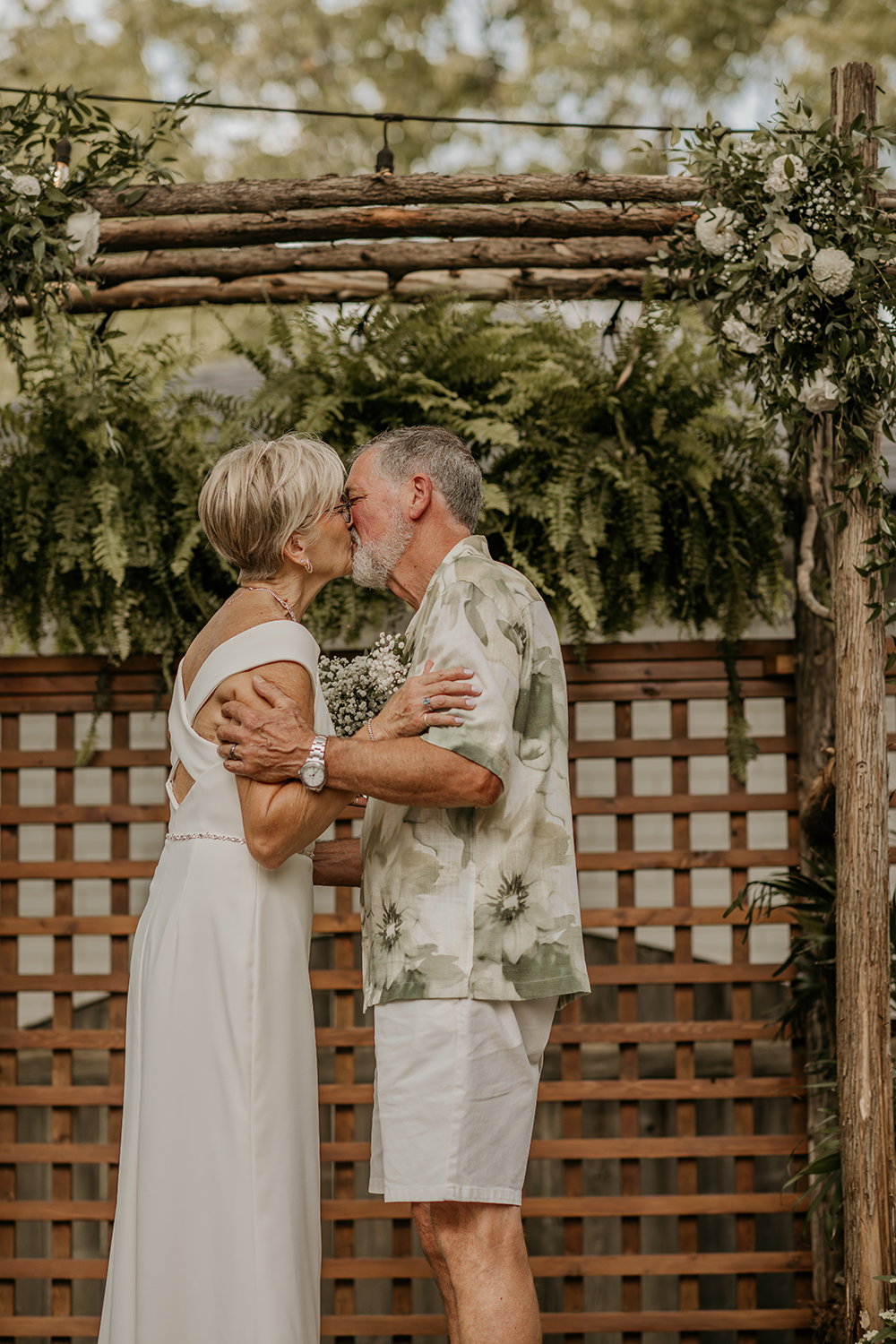 A bride and groom are kissing during their wedding ceremony under a wooden arch.