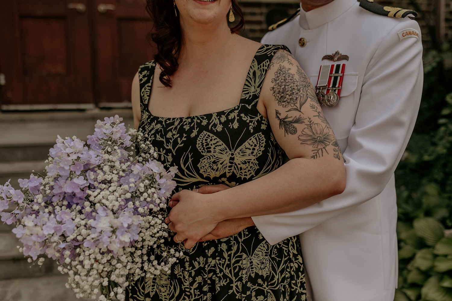 A man in a white uniform is hugging a woman in a black dress who is holding a bouquet of purple flowers.