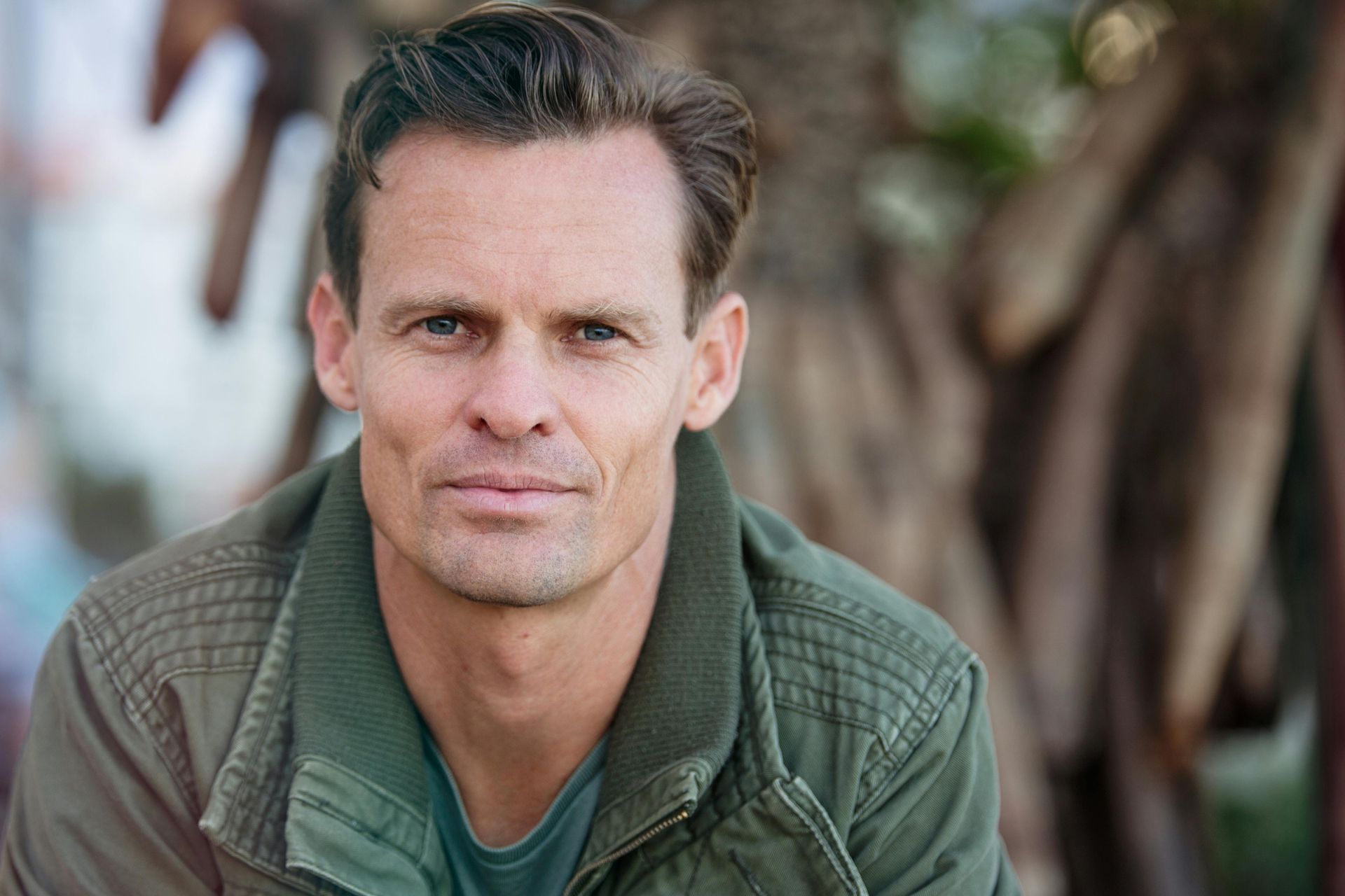 A man in a green jacket is sitting in front of a tree and looking at the camera.