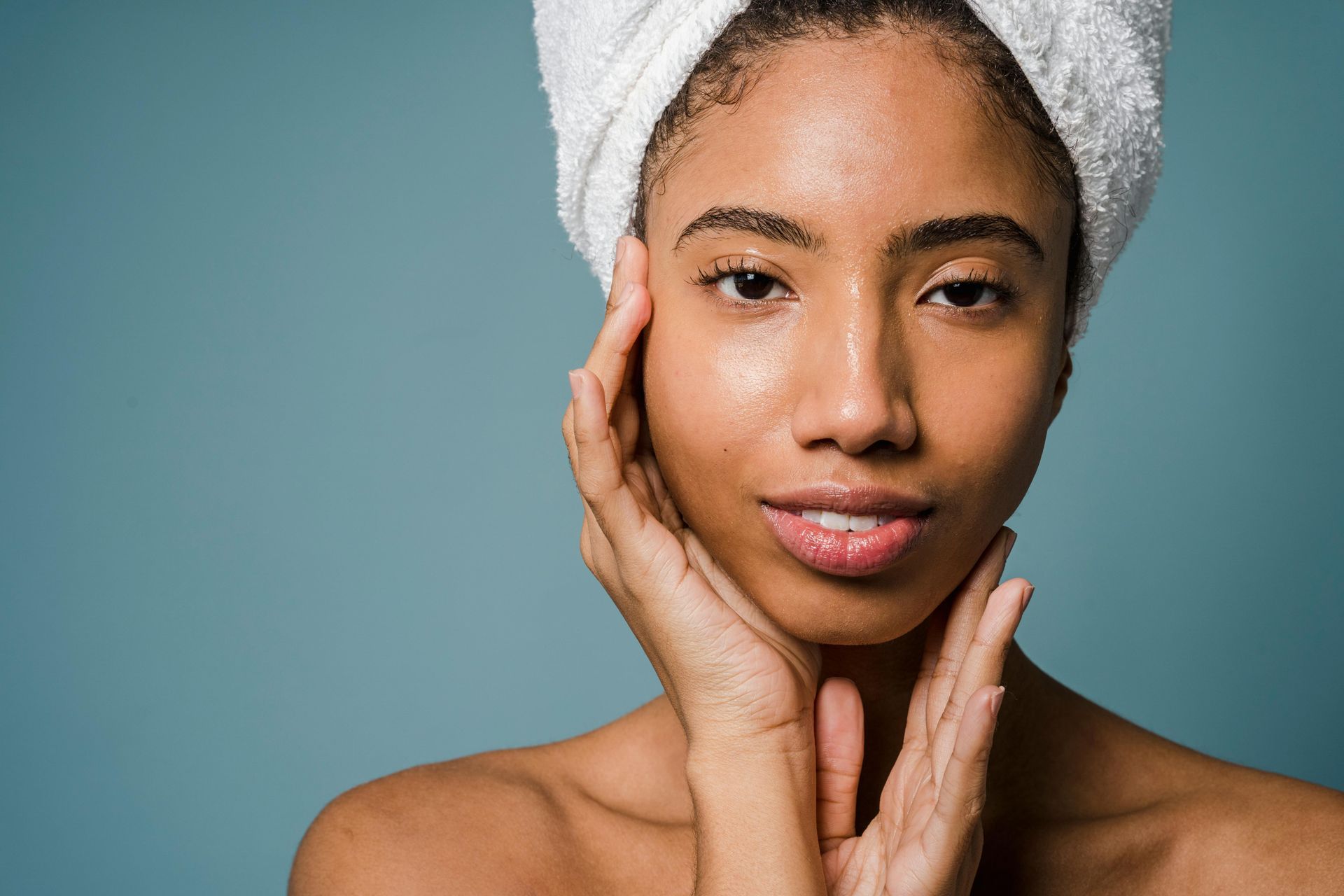A woman with a towel wrapped around her head touches her face.