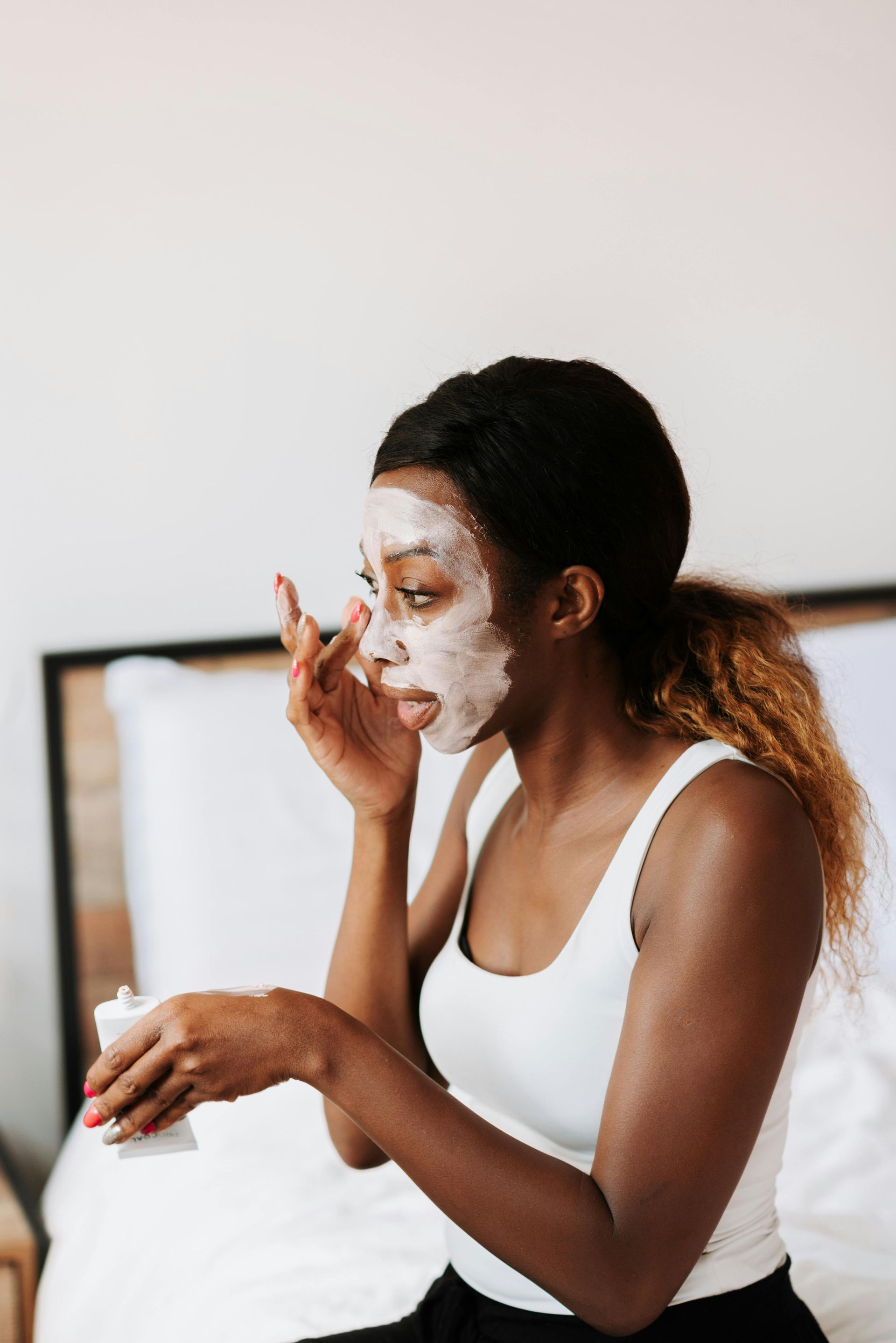 A woman is sitting on a bed applying a face mask.