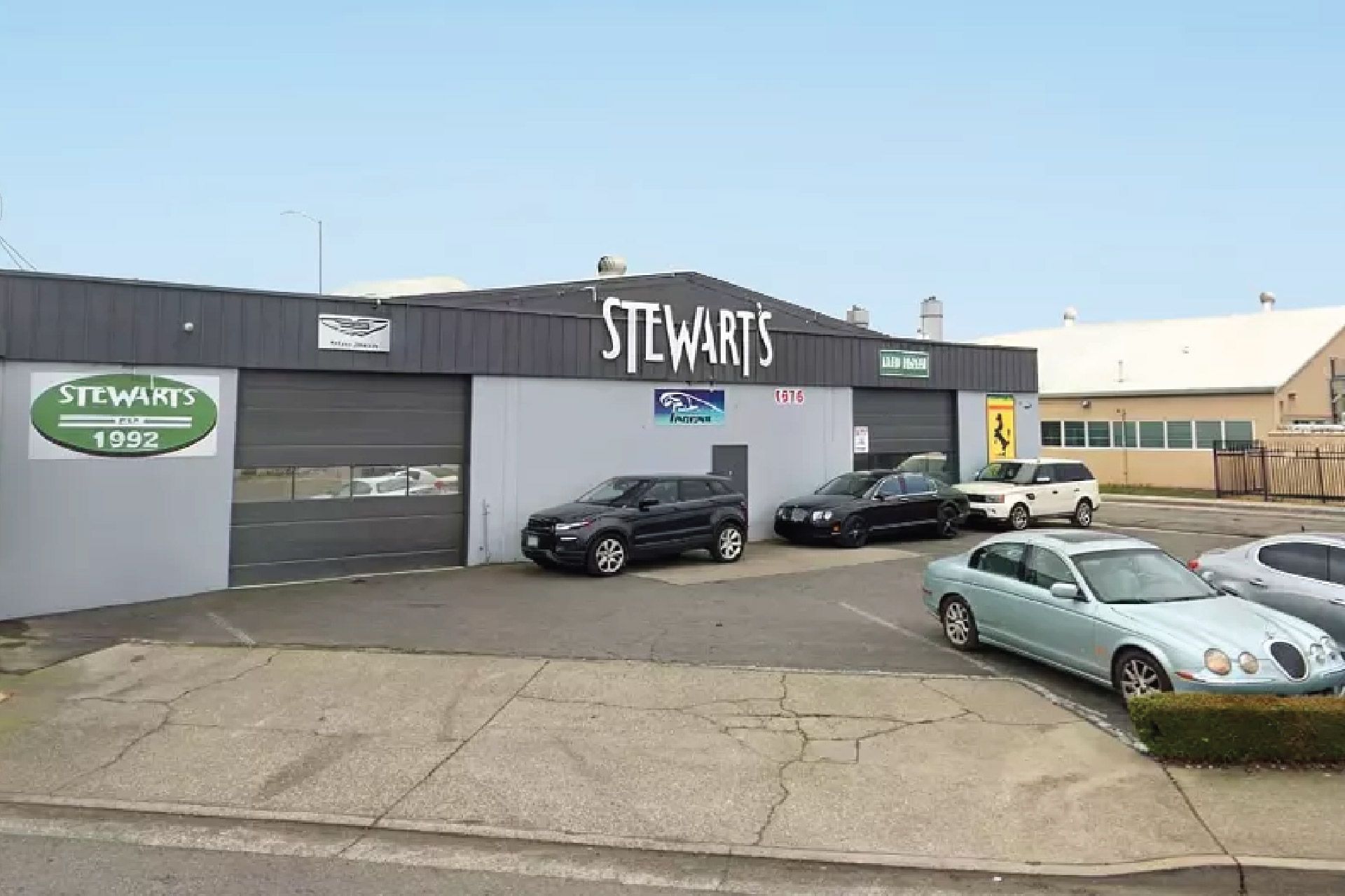 Stewart's auto repair shop with several cars parked outside. Gray building, green sign, blue sky.