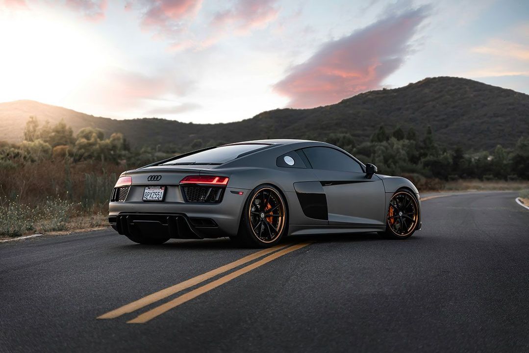 Dark gray Audi R8 sports car on a road, mountains in the background, sunset colors in the sky.