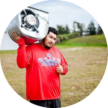 Man in red shirt holding a large metal tank on his shoulder, giving a thumbs up outdoors.