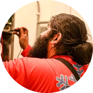Man with long beard in red shirt, working on electrical panel, indoor setting.