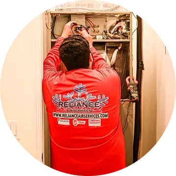 HVAC technician in red shirt repairing furnace, holding tools. White wall.
