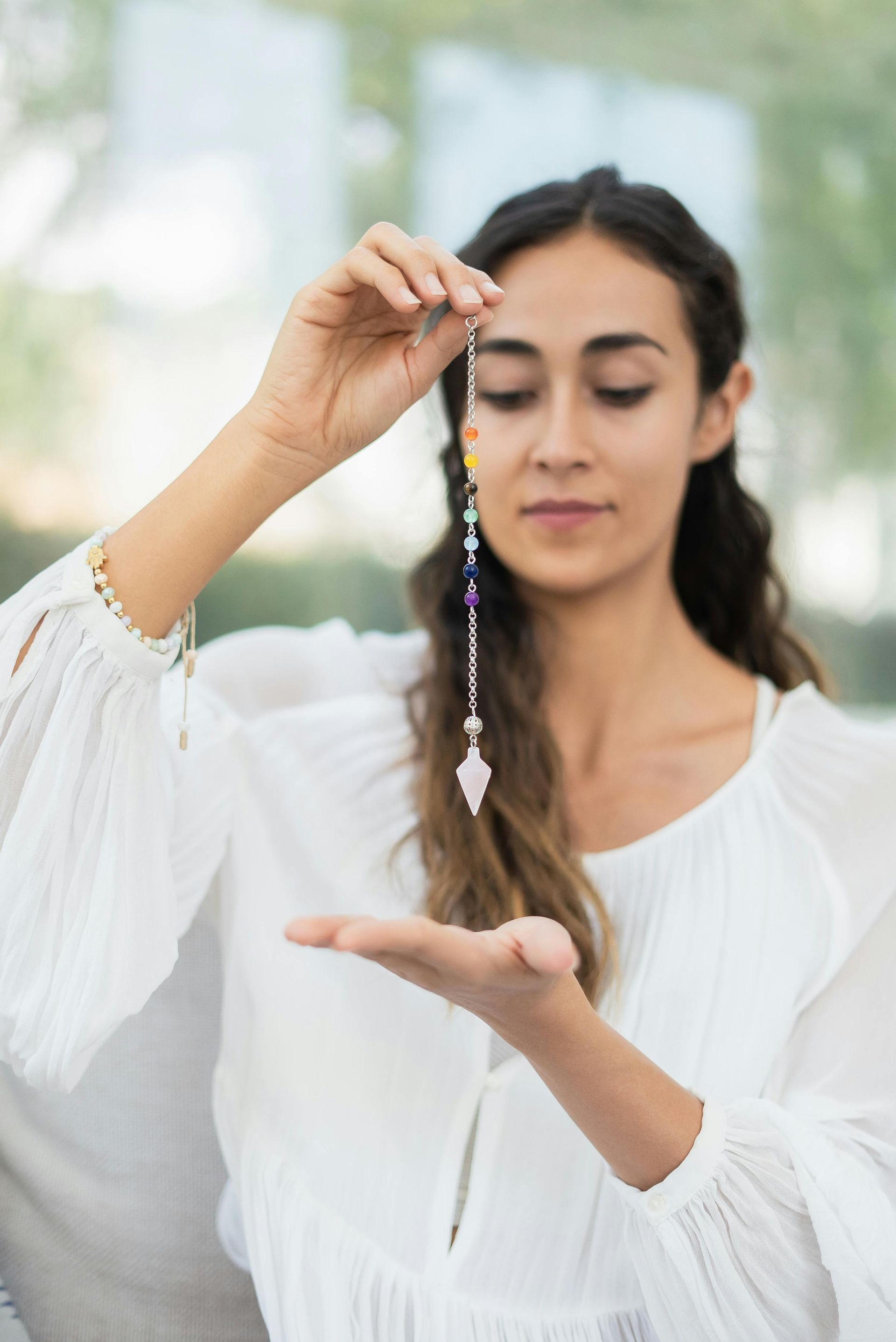 Woman holding a pendulum with chakra beads, gazing at it with a focused expression.