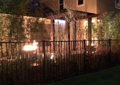 Nighttime backyard scene with bamboo, metal fence, and illuminated water feature.