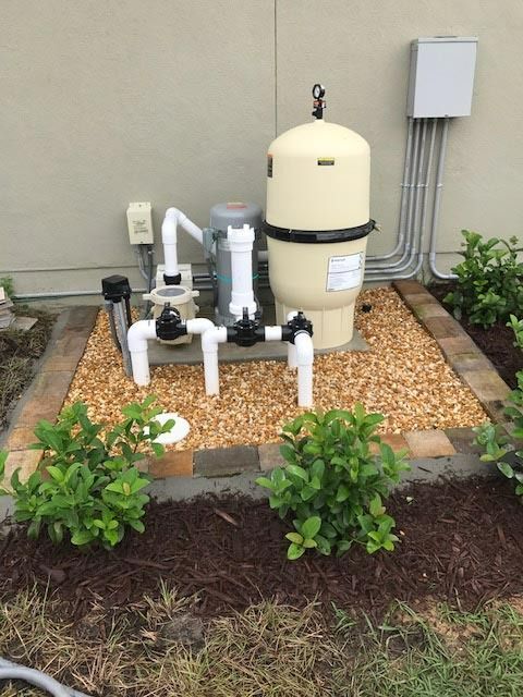 Pool equipment setup: Beige filter, pipes, electrical box, and small bushes on gravel bed.