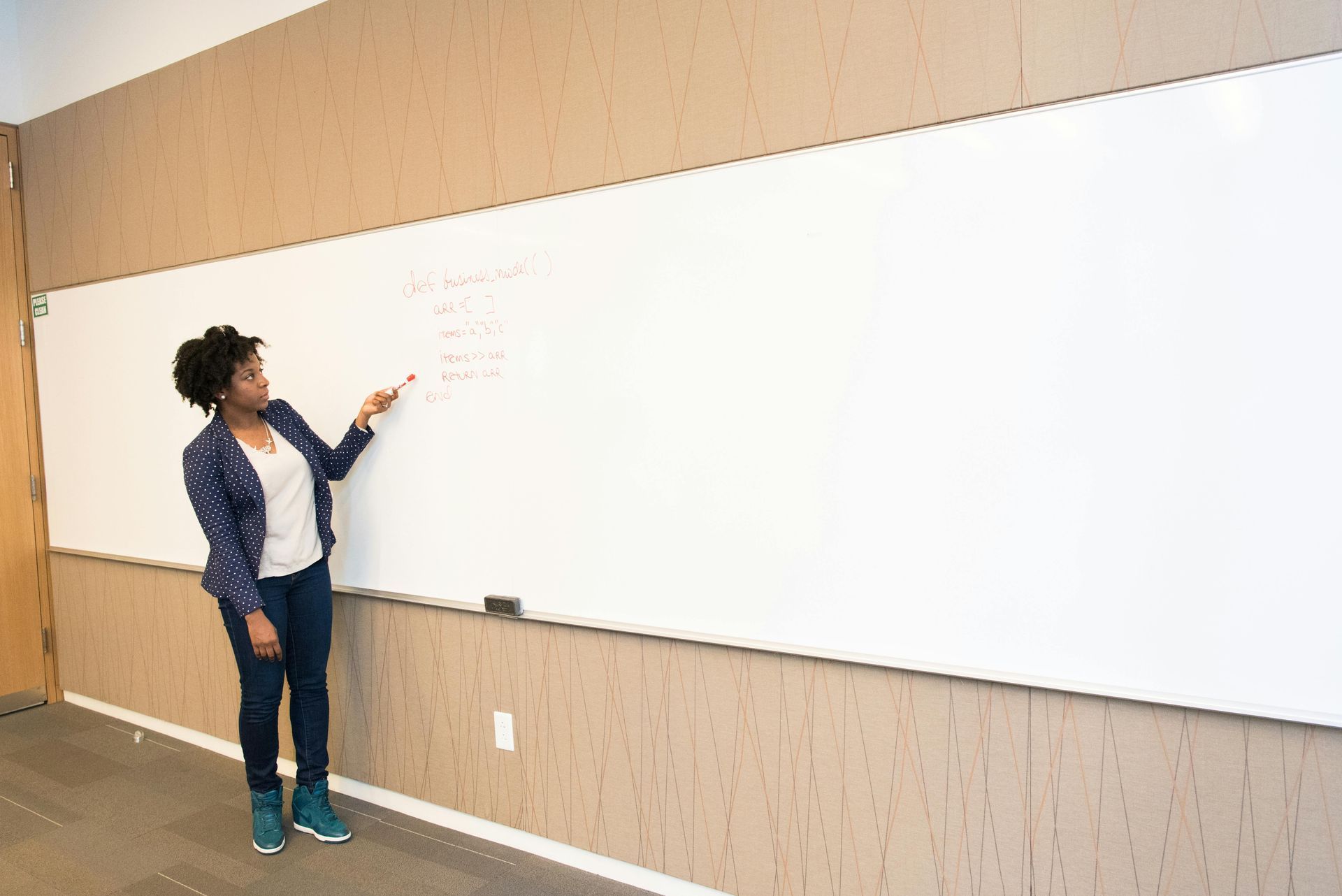 Woman with curly hair points to a whiteboard with writing in a light-toned room.