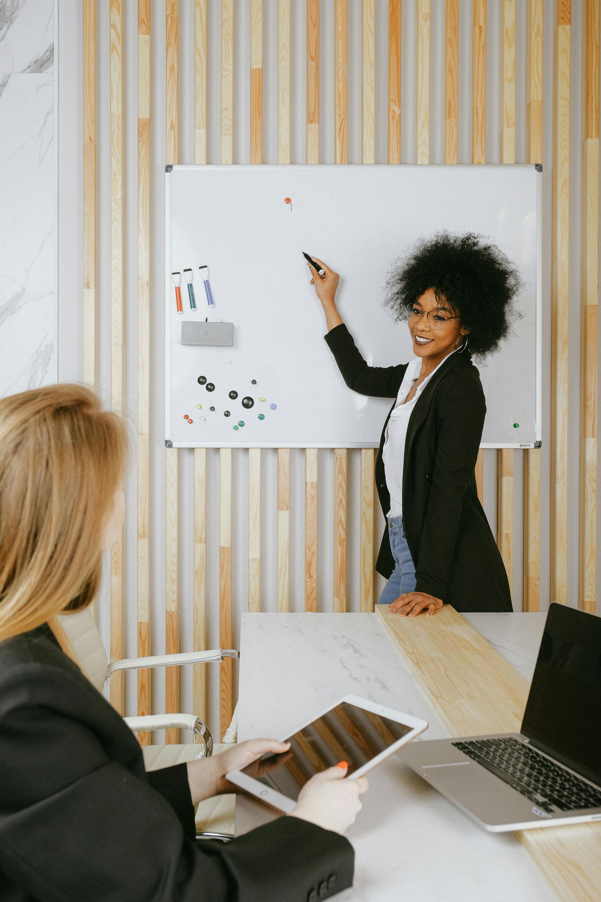 Woman presenting at a whiteboard, pointing with a marker, while a colleague uses a tablet and laptop.