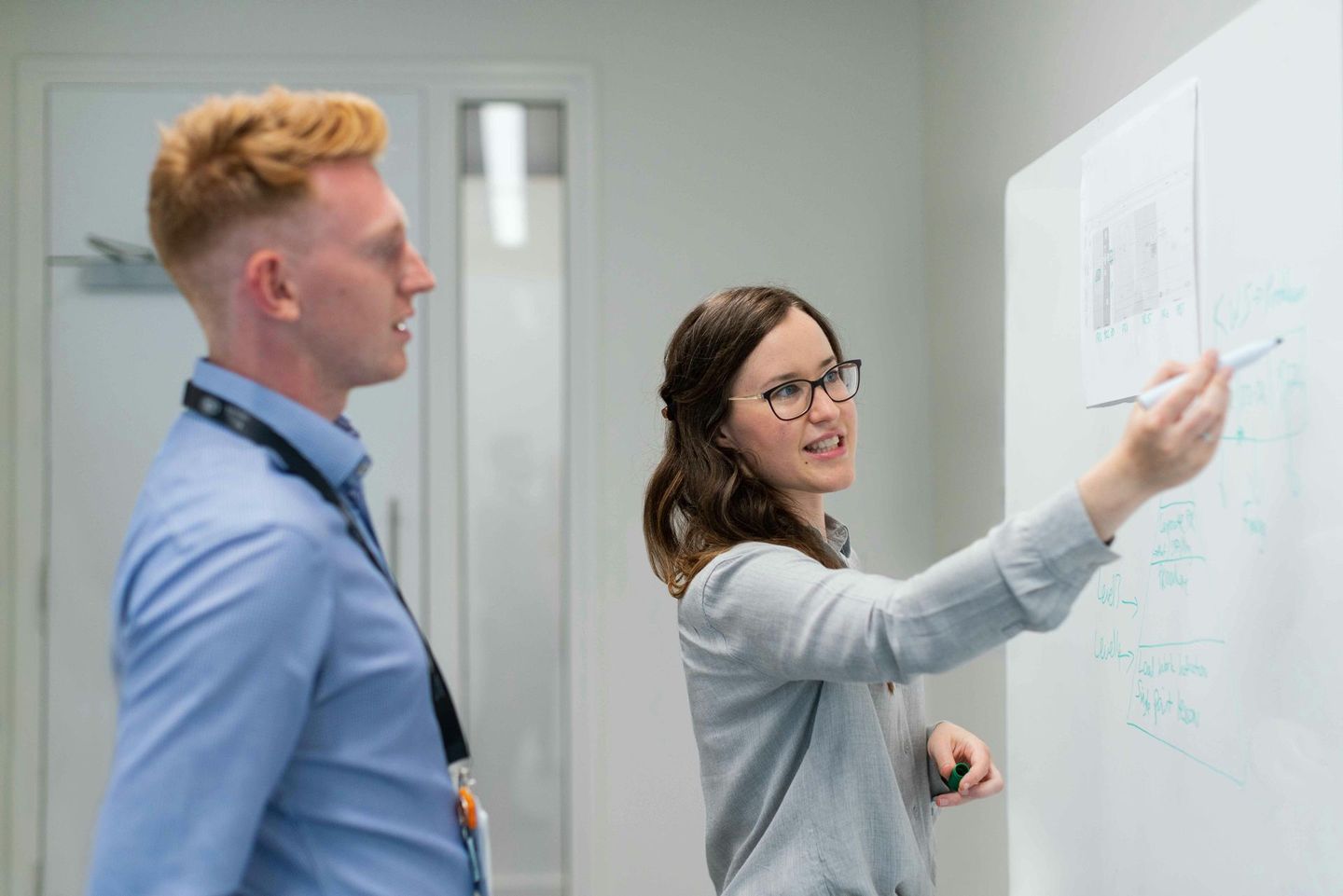 A woman in glasses explains a diagram on a whiteboard to a man in a blue shirt.