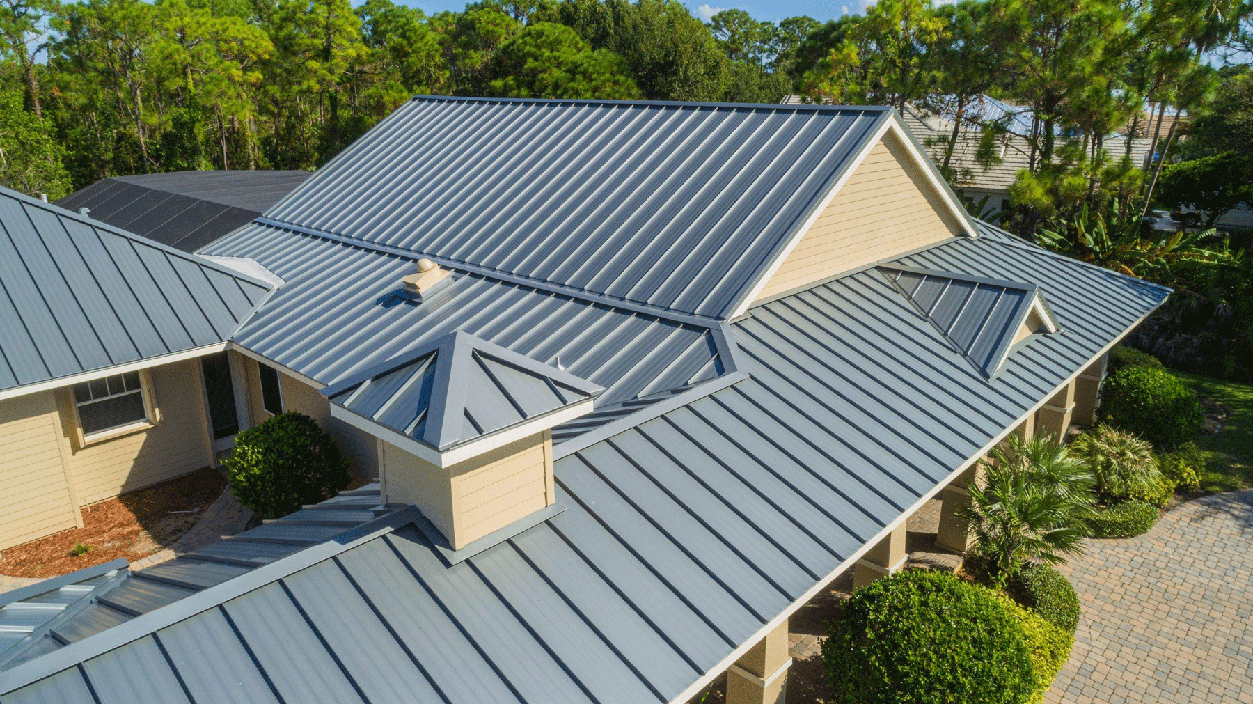 Modern gray metal roof on a large house with dormers, surrounded by green trees and shrubs.