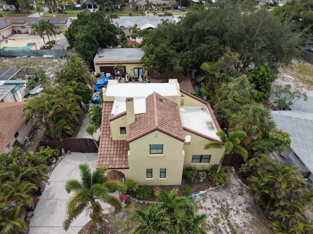 Aerial view of a beige house with a red tile roof surrounded by trees and neighboring homes.