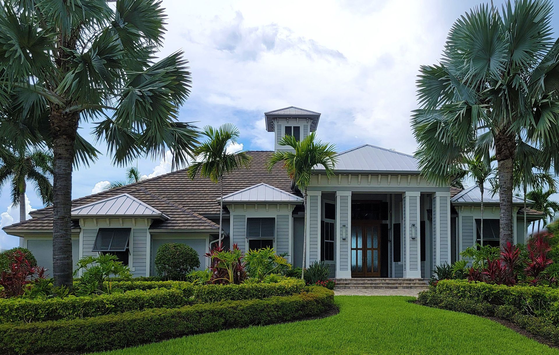 Modern tropical house with palm trees, manicured lawn, and gray roof under a cloudy sky