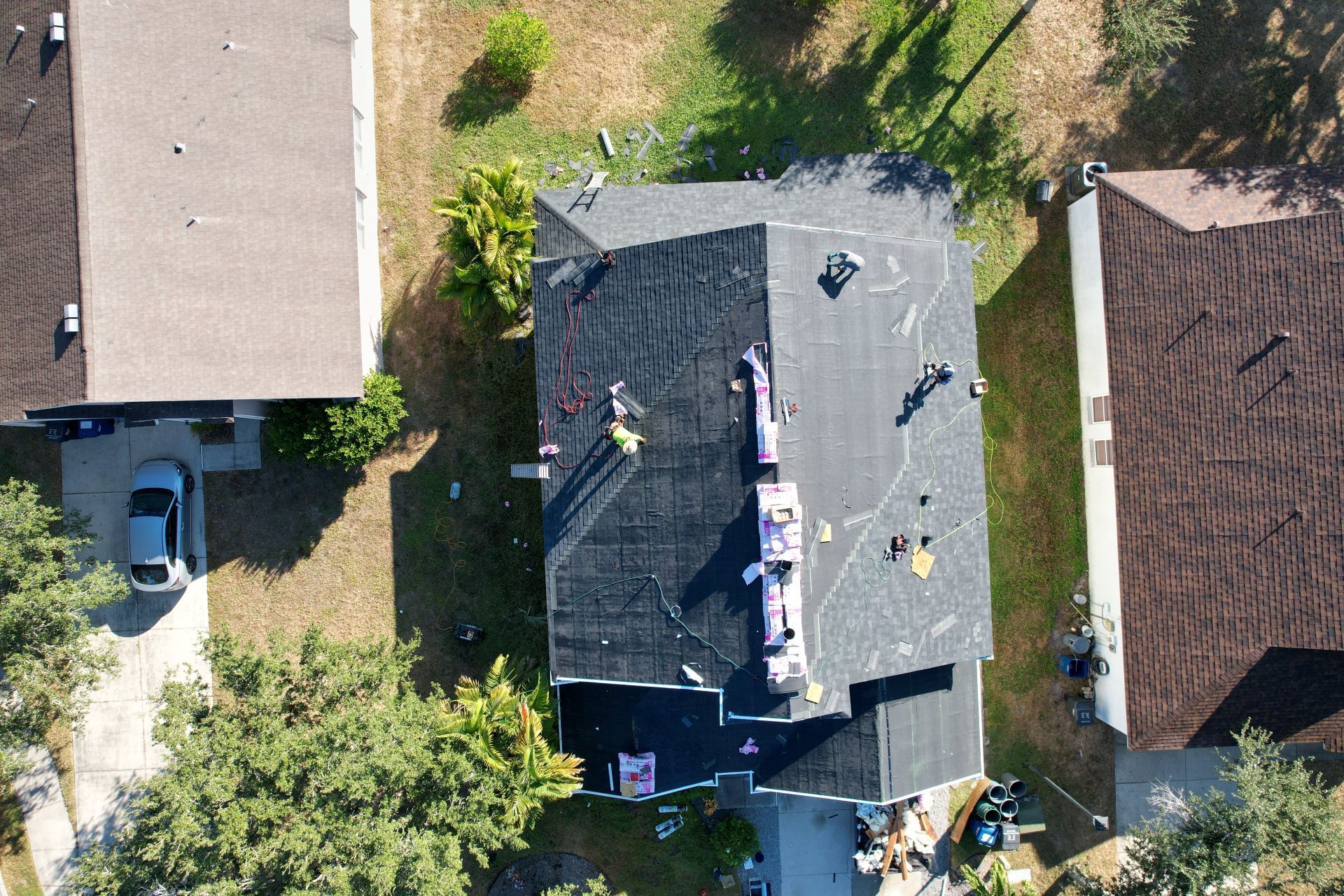 Aerial view of a house roof with workers on it, surrounded by trees and neighboring homes.