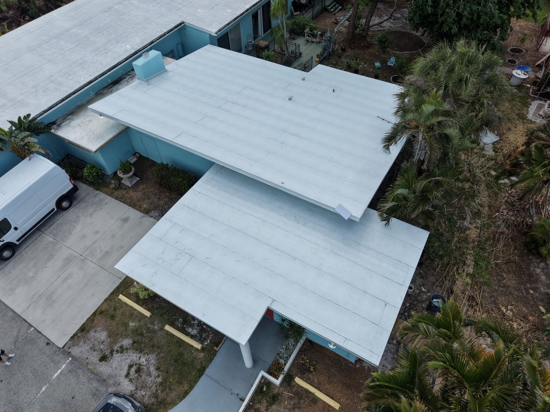 Aerial view of a white-roofed house with a carport, driveway, and surrounding greenery